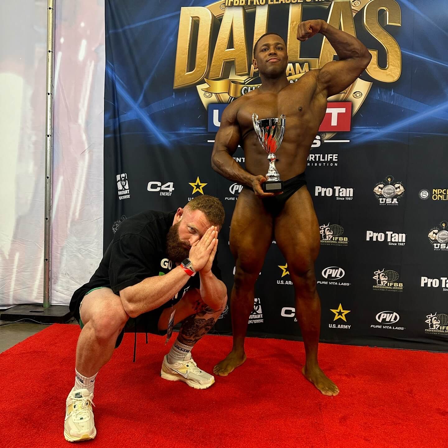 Personal Trainer Marlin Foy kneels in a victory pose by a client who is flexing and holding a trophy
